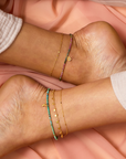 Light-skinned bare feet rest on soft peach fabric, adorned with the 9" Turquoise Moon Anklet plus a 1" extender chain, featuring delicate gold charms, beads, and a turquoise moon detail.
