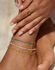 A close-up of manicured hands resting on an ankle adorned with the Joyful Anklet, featuring an 18k gold plate oval cable chain and beaded accents. A gold ring graces one finger, and the skin has a soft, natural tone.
