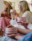 Four women gather for The Soul of Tea: A Two Day Retreat in Ojai, CA, each holding a pink ceramic bowl. Their softly blurred faces place emphasis on the bowls and their hands, while their pastel attire enhances the serene and tranquil ambiance of the tea ceremony.