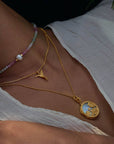 A close-up of a woman in a white textured blouse wears three layered necklaces, including the Loving Devotion Hummingbird Necklace 16", a multicolored beaded strand, and a large round gold locket with an embossed design.