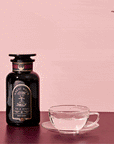 A black bottle labeled "Ruby Moon™ Herbal Tea Sachets" sits on a pink surface beside a transparent glass teacup, as caffeine-free herbal tea is poured into the cup against a pink background.