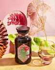 A dark glass bottle labeled "Queen of Kyoto" and a round glass sit on a surface, surrounded by Jasmine Blossoms, pink and gold decor, green leaves, and a reflective fan against a softly vibrant pink and gold background.