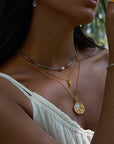 A woman wearing a white off-shoulder top is shown from the shoulders up, accessorized with layered gold necklaces including the Loving Devotion Hummingbird Necklace 16", plus a beaded bracelet, gold ring, and drop earring in soft natural light.