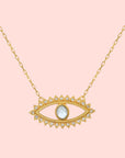 The Eye Blue Topaz Necklace features a gold pendant in an eye shape, accented with clear gemstones and a central blue topaz stone, inspired by the classic evil eye design.