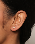 Close-up of a woman's ear wearing White Topaz Lotus Studs—delicate earrings crafted with 18kt gold plate and three sparkling stones—set against a neutral background.
