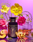 A display featuring the Monthly Magic Tea Journey Subscription—an ornate tea tin, glass teapot, rare botanicals, loose tea leaves, peaches, orchids, roses, and yellow poppies—evokes a goddess ritual on a vibrant pink background.