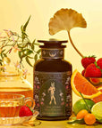Freya: The Radiant tea set features a glass teapot and wellness tea cup beside a dark floral canister, surrounded by strawberries, orange slices, lime, kumquats, jasmine flowers, and a gold ginkgo leaf on a yellow background.