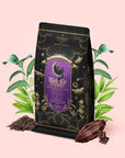 A bag of Black Cat Brownie: Decadent Chocolate Puerh Blend with Vanilla & Hazelnut, featuring a purple and gold label, is surrounded by green leaves, a cocoa pod, and pieces of chocolate—this dessert-inspired black tea blend is displayed on a light pink background.