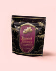 A black and gold tea package labeled “Haunted Hibiscus: Tart Berry Herbal Blend with Warm Spices,” adorned with moth and flower illustrations and celestial designs, rests on a pale pink background. This enchanting caffeine-free tea blends tart berries and warm spices for a truly mystical experience.