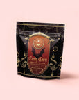 A black and gold package of Candy Corn: Herbal Fall Tea with Vanilla & Caramel, adorned with mystical designs, a bat, and plant illustrations, sits against a pale pink background. This caffeine-free blend is the ideal Halloween treat for anyone seeking the sweet taste of candy corn in every cozy sip.