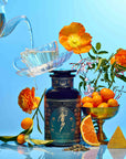 A teapot pours tea into a floating teacup above a “Magic Hour” jar, evoking goddess rituals. Surrounded by orange flowers, kumquats, an orange slice, leaves, and a golden pyramid—all showcase the Monthly Magic Tea Journey Subscription.