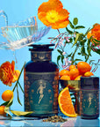A teacup is being filled with tea beside two "Monthly Magic Tea Journey Subscription" containers—ideal for a goddess ritual—surrounded by orange flowers, kumquats, an orange slice, and loose tea leaves against a vibrant blue background.