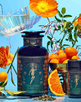Hathor of Joy: Honey Nectar Herbal Tea is poured into a teacup beside a large ornate tea jar, small tin, kumquats, orange slice, tea leaves, and orange flowers on a vibrant blue background.