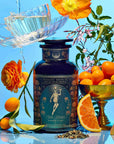A bottle of Hathor of Joy: Honey Nectar Herbal Tea is surrounded by sliced citrus fruits, orange kumquats in a gold bowl, orange flowers, and a teacup with water splashing out against a vibrant blue background.