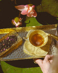 A hand pours tea from the Monthly Magic Tea Journey Subscription into a patterned teacup on a gold tray beside loose tea leaves, with pink lotus flowers and green lily pads floating in water in the background—an invitation to a goddess ritual that honors the Divine Feminine with every soothing sip.