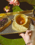 A hand pours Lakshmi in Abundance: Rose Saffron Black Tea for Good Fortune & Radiance into a yellow floral teacup set on a gold tray with loose tea leaves and petals, surrounded by blooming lotus flowers and green lily pads.
