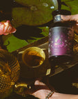 A hand holds a jar of Lakshmi in Abundance: Rose Saffron Black Tea for Good Fortune & Radiance above a brass tray with a teapot and cup. Pink lotus flowers and lily pads grace the background, setting the scene for savoring this antioxidant-rich, luxury black tea.
