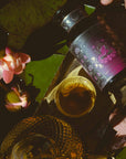 A hand holds a jar of Monthly Magic Tea Journey Subscription tea with a purple floral label next to a teacup, teapot, water lily flowers, and lily pads, all bathed in warm sunlight—perfect for a goddess ritual celebrating the Divine Feminine.