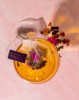 A glass of herbal tea is shown with a tea bag steeping inside. The tag on the tea bag reads "Magic Hour." Several loose tea leaves and flower petals are scattered next to the glass on a pink surface, reminiscent of a serene tea ceremony. The warm, amber hues of the tea come from the Magic Hour Starter Kit by Magic Hour.