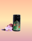 A Moonlight in Marrakesh: Spiced Mint Green Tea case pack by Magic Hour, with its black cylindrical container and decorative green and gold design, stands on a white surface. Surrounding it are pink and purple orchids, lavender flowers, and hints of Moroccan mint tea, adding a touch of nature and color to the scene.