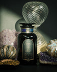 A dark glass jar labeled "Moringa Moon Herbal Tea" stands on a table with amethyst crystals, gourd-shaped glass ornaments, and a spoonful of loose caffeine-free tea, illuminated by dramatic lighting and shadows.