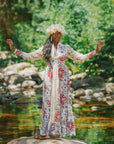 A person in a floral dress and flower crown stands with arms raised on a rock by a stream, surrounded by greenery. At their feet rests a teapot of Oshun’s Nectar: Honeyed Herbal Tea for Joy & Radiance and a jar.