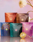 The Queen's Sampler Set features five vibrant tea packages with gold designs in a row. In front, a gold tea infuser and crescent moon charm complete this charming wellness tea set, accented by soft pink flowers on a pastel background.
