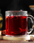A steaming glass mug from the Heart Health Tea Sampler Set, filled with vibrant red hibiscus tea known for its cardiovascular wellness benefits, is garnished with dried hibiscus petals and rests on a wooden coaster. The softly blurred background evokes a cozy, warm setting perfect for enjoying herbal blends.