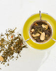 A metal tea infuser filled with Magic Hour organic wellness tea and sliced nuts from The Best of Magic Hour Sampler Set with Tea in Hand Teapot rests in a cup of green tea, with loose leaves scattered on the surface nearby.