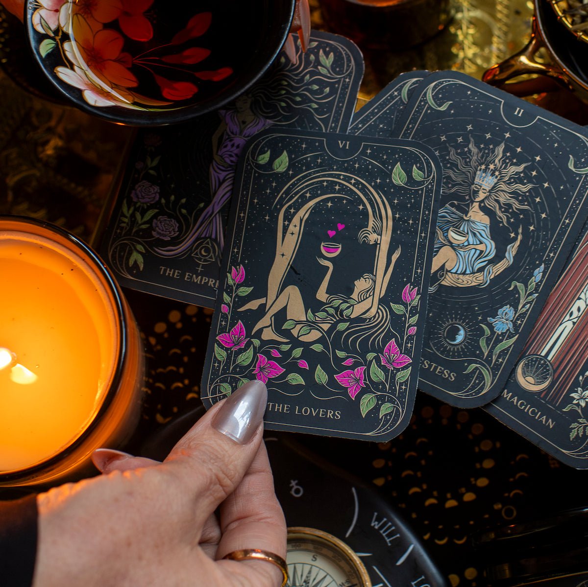 A hand with silver-painted nails holds "The Lovers" tarot card over a lit candle and several other tarot cards, including "The Empress," "The High Priestess," and "The Magician." The scene showcases elements from The Lovers Box, which includes a floral teacup filled with the Lovers tea blend and a compass.