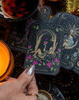 A hand with silver-painted nails holds "The Lovers" tarot card over a lit candle and several other tarot cards, including "The Empress," "The High Priestess," and "The Magician." The scene showcases elements from The Lovers Box, which includes a floral teacup filled with the Lovers tea blend and a compass.