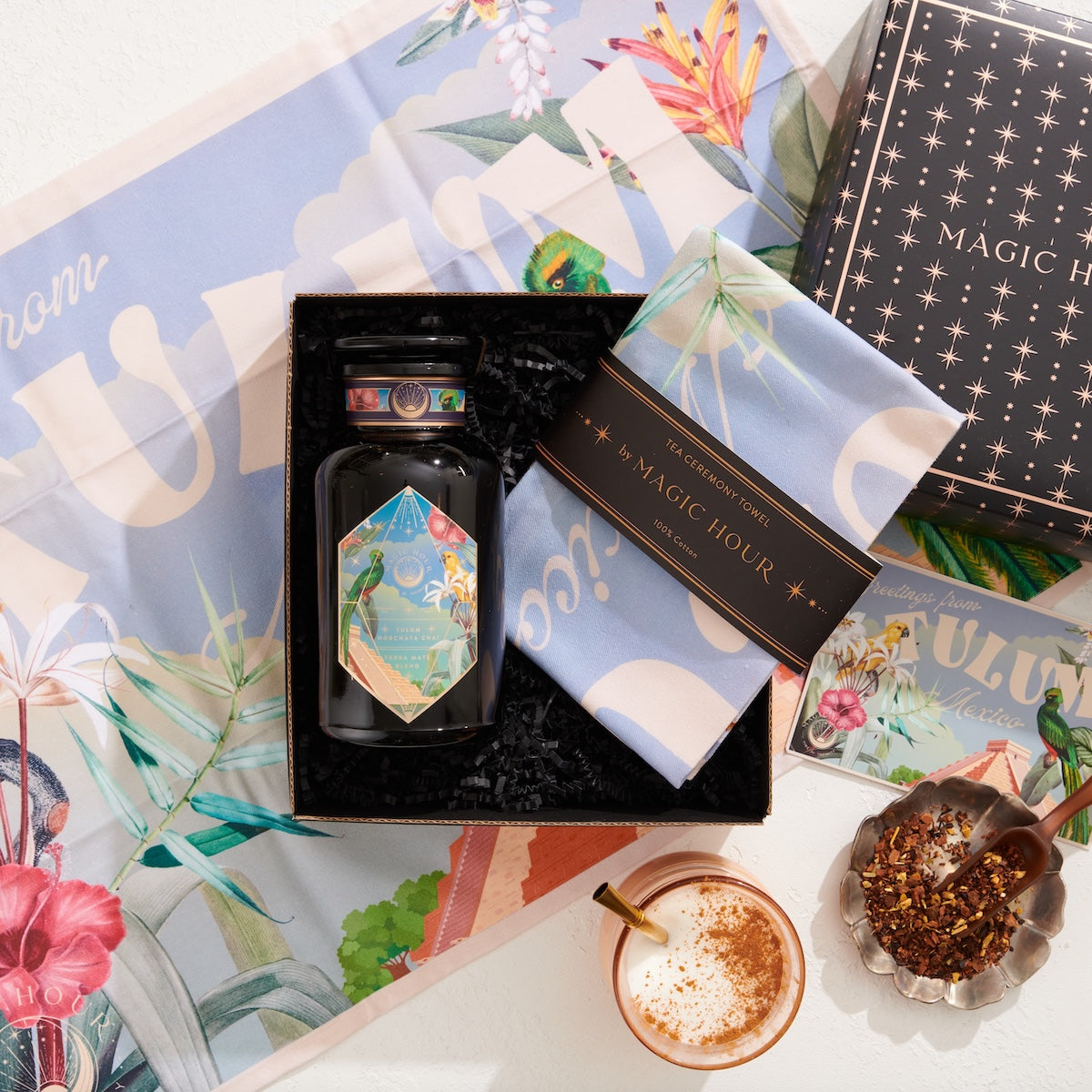 The Travel to Tulum Gift Set includes a tea jar, pastel cloth, and "Magic Hour" box. Experience the unique Tulum Horchata Chai with its loose leaves on floral-themed paper against a vibrant backdrop for an enchanting tea moment.