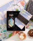 The Travel to Tulum Gift Set includes a tea jar, pastel cloth, and "Magic Hour" box. Experience the unique Tulum Horchata Chai with its loose leaves on floral-themed paper against a vibrant backdrop for an enchanting tea moment.