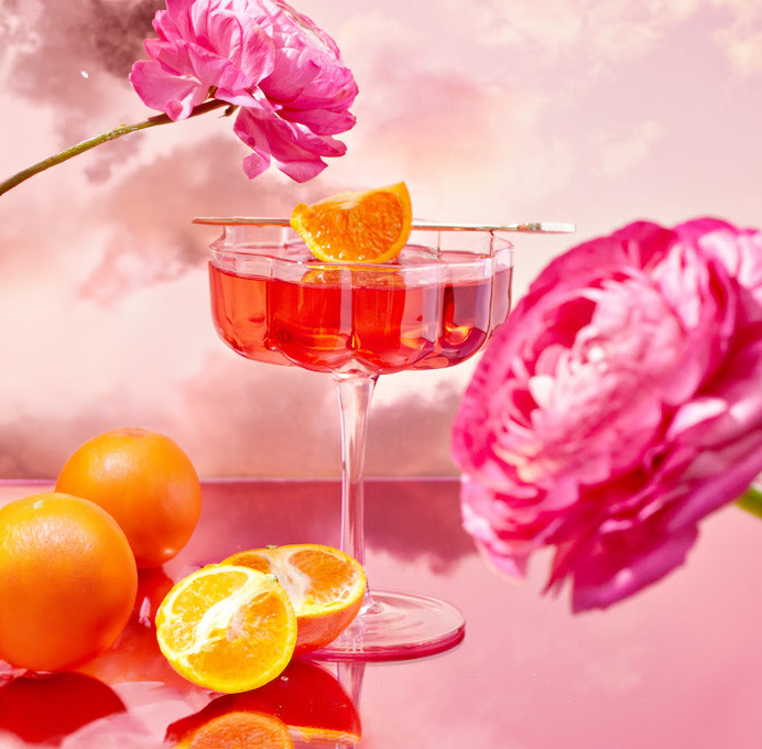A tea from the Wanderlust Sampler Tea Set in a delicate cup garnished with an orange slice sits on a glossy surface surrounded by whole and halved oranges. Pink flowers and a cloudy, pastel sky form a vibrant and dreamy backdrop.