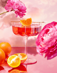 A tea from the Wanderlust Sampler Tea Set in a delicate cup garnished with an orange slice sits on a glossy surface surrounded by whole and halved oranges. Pink flowers and a cloudy, pastel sky form a vibrant and dreamy backdrop.