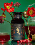 A tin of "Persephone's Potion: Pomegranate & Jasmine Green Tea" sits among red flowers, a glass teapot with pomegranate tea, a glass cup, a gold infuser, and cranberries on a vibrant green backdrop.