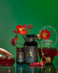 A hand reaches for a glass teacup filled with Persephone's Potion: Pomegranate & Jasmine Green Tea, surrounded by ornate packaging, a teapot, red flowers, cranberries, and a tea strainer on a vibrant green background.