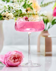 A pink cocktail in a coupe glass with a cherry, surrounded by splashes and a pink rose, is set against blurred vases of flowers. The scene evokes the bright elegance of Diamond - Champagne & Strawberry Jasmine White Tea for Beautiful Skin.