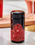 A dark glass jar of Carnelian : Caffeine-Free Apricot Amaretto Tea with a red geometric label rests on a marble surface, surrounded by blurred glassware and a wooden tray in the background.