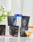 A display of five elegant tea packages with floral designs, including Magic Hour's The Astrology Collection - Luxe Sampler Sips, arranged around three different tea glasses containing colorful beverages. A vase with white flowers is in the background, adding to the serene and sophisticated atmosphere.