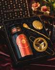 A Season of Abundance Gift Set contains a black tea canister with a celestial design labeled "Magic Hour," a gold tea infuser, and a golden teaspoon. The set is adorned with flowers and includes loose leaf organic tea, all beautifully presented in a matching themed box featuring stars and "Magic Hour" text.