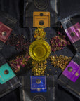 A circular arrangement of six colorful tea packages surrounds a glass filled with tea. The packages, each of a different color, have intricate designs on them. Various loose tea ingredients are scattered in the center around the glass on a dark background, completing the Self-Guided 8 Day Ceremonial Tea Cleanse Kit.