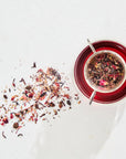 A glass cup filled with Immune Boosting Teas Sampler Set by Magic Hour and a tea strainer containing loose leaf tea rest on a white surface. A small pile of scattered tea leaves is visible beside the cup. The organic tea has a deep red color, and the scene is brightly lit.