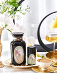 Elegant display featuring two black jars with pastel labels, a tall glass of golden liquid from Pearl Tea of Beauty, Clarity, and a Sense of Peace by Magic Hour, a gold saucer with a white cup, and delicate flowers in the background, including orchids. A small statue adds a touch of classical charm to the scene.