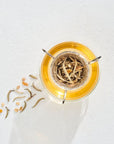 A glass of golden-colored Magic Hour The Astrology Collection - Luxe Sampler Sips viewed from above, with a strainer containing loose leaf tea and other organic ingredients submerged in the center. Some tea leaves and small pieces are scattered on the textured white surface next to the glass.