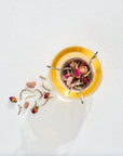 Top view of a glass cup with a metal infuser holding Magic Hour's The Astrology Collection - Luxe Sampler Sips, including rose buds and leaves, over a white textured background. Some loose leaf tea is scattered around the cup, adding to the aesthetic. The cup contains a light-colored organic tea.