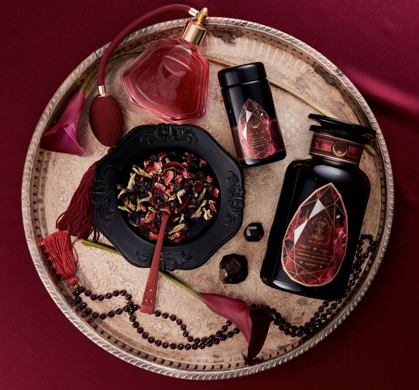 A decorative tray holds a bowl of dried flowers with a wooden spoon, a red perfume bottle, a black round bottle with a red ornate label, and black beads. A cylindrical black container of Magic Hour Garnet Gemstone Box completes the scene. The background and some flower petals are deep red, creating a luxurious ambiance.