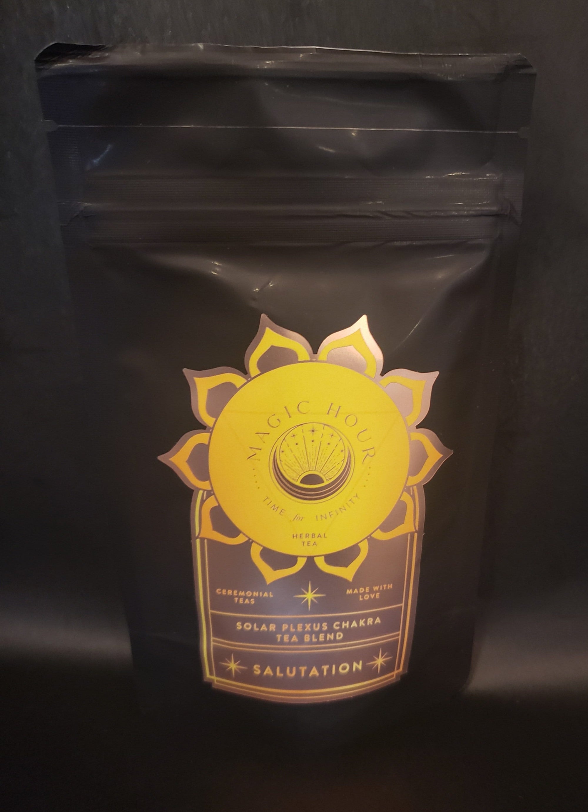 A black resealable pouch of Salutation™ Tea for the Solar Plexus Chakra is displayed against a dark background. The packaging features a golden geometric design with a sun motif and details "Solar Plexus Chakra Tea Blend - Salutation" in elegant gold text, highlighting its organic ingredients for optimal chakra balance.