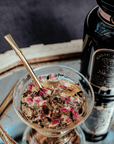 A jar labeled "Magic Hour" stands next to a glass filled with a mixture of herbs and dried rose petals, complemented by an Alibaba Sips Tea Straw & Strainer Bombilla resting inside. Both items are set on a reflective surface, beautifully capturing the light.