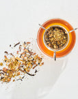 A glass teapot filled with orange-hued Magic Hour Immune Boosting Teas Sampler Set is photographed from above. Loose Leaf Tea and herbs are scattered beside the pot on a white surface, creating a colorful mix of ingredients. The image is bright with natural light casting soft shadows.